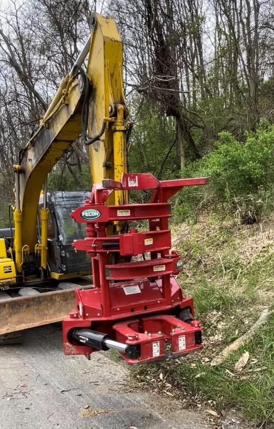 An image showing a closer view of a red tree shear attachment as an extension of a yellow PennDOT construction and maintenance vehicle in a woodland setting.